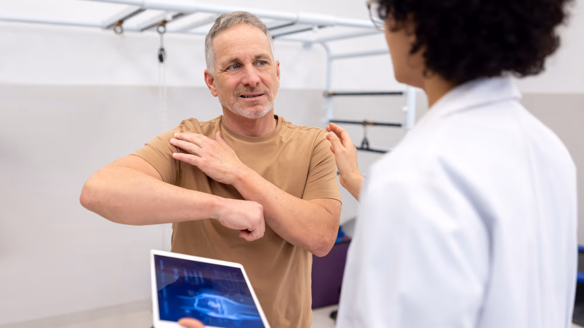 Man in tan shirt showing shoulder pain to healthcare professional holding a tablet with an X-ray image.