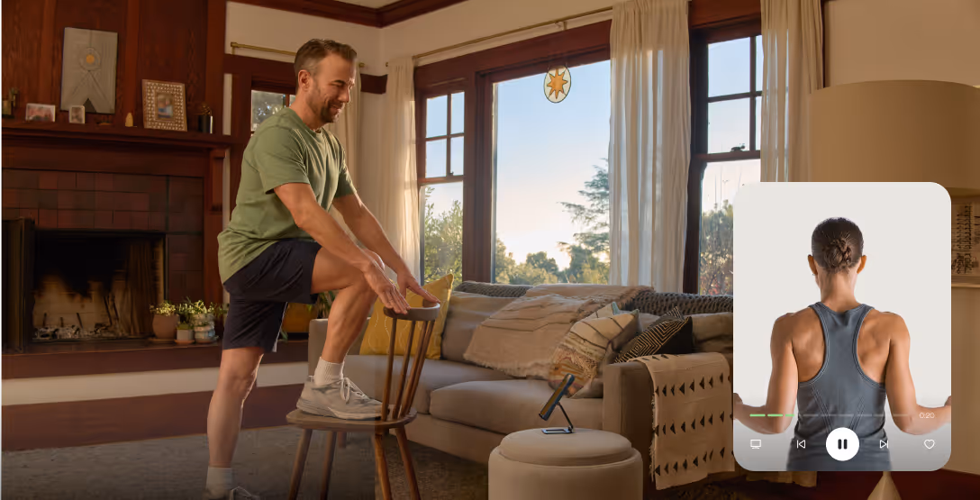 Man in a green shirt and black shorts stretching one leg on a chair in a cozy living room, following a workout video of a woman shown on a nearby screen.