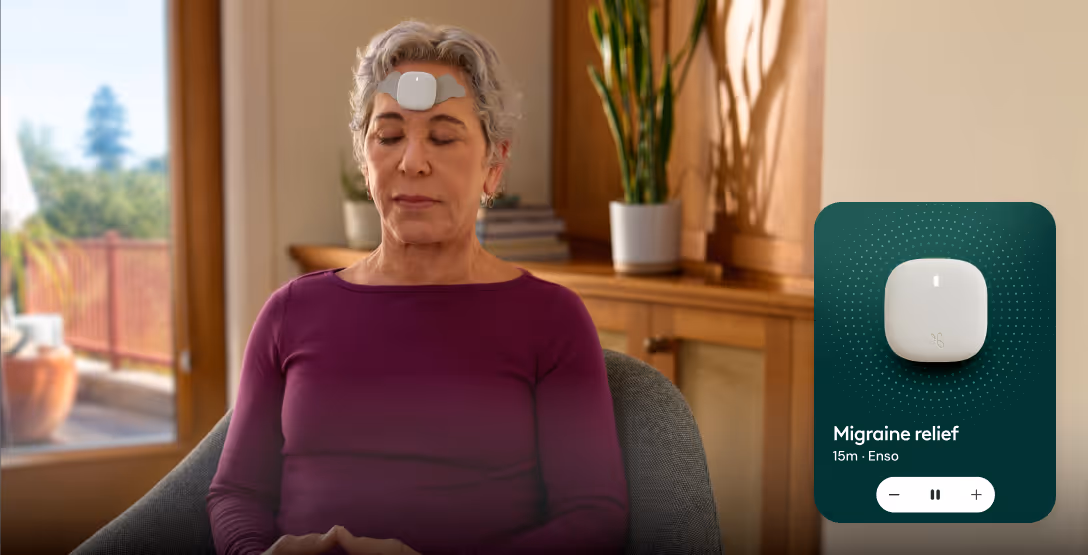 Older woman with eyes closed wearing a white migraine relief device on her forehead, sitting in a chair with a plant and wooden cabinet in the background.