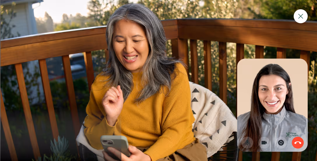 Woman in mustard sweater smiling while video chatting on smartphone with another woman wearing a Hinge Health jacket.