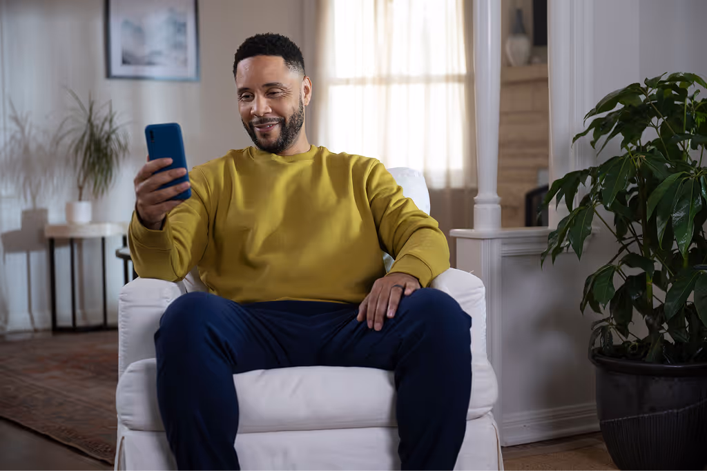 Smiling man in yellow sweater sitting on a white armchair looking at his phone in a cozy living room.