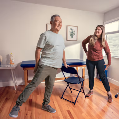 Senior man stretching his leg while holding a chair in a room with a woman instructor watching and smiling.