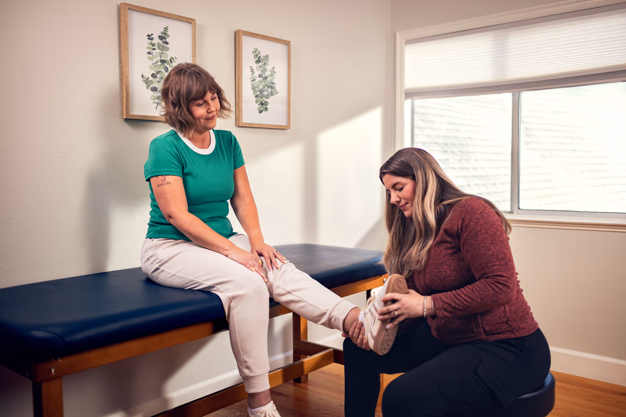 A woman in a green shirt sits on an examination table while another woman in a maroon jacket examines her foot in a medical office.