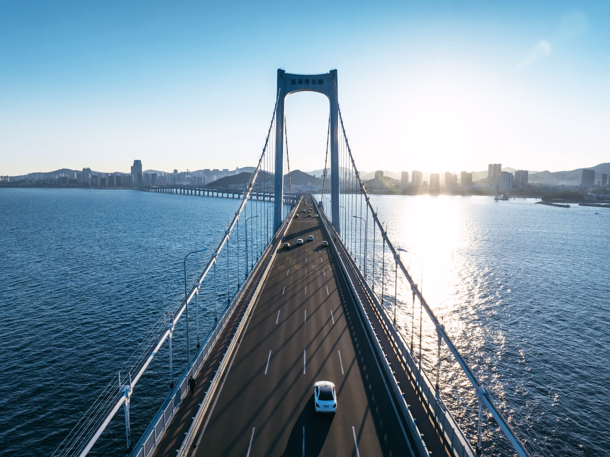 Aerial view of cars traveling across a suspension bridge over the water, with a city skyline in the distance.