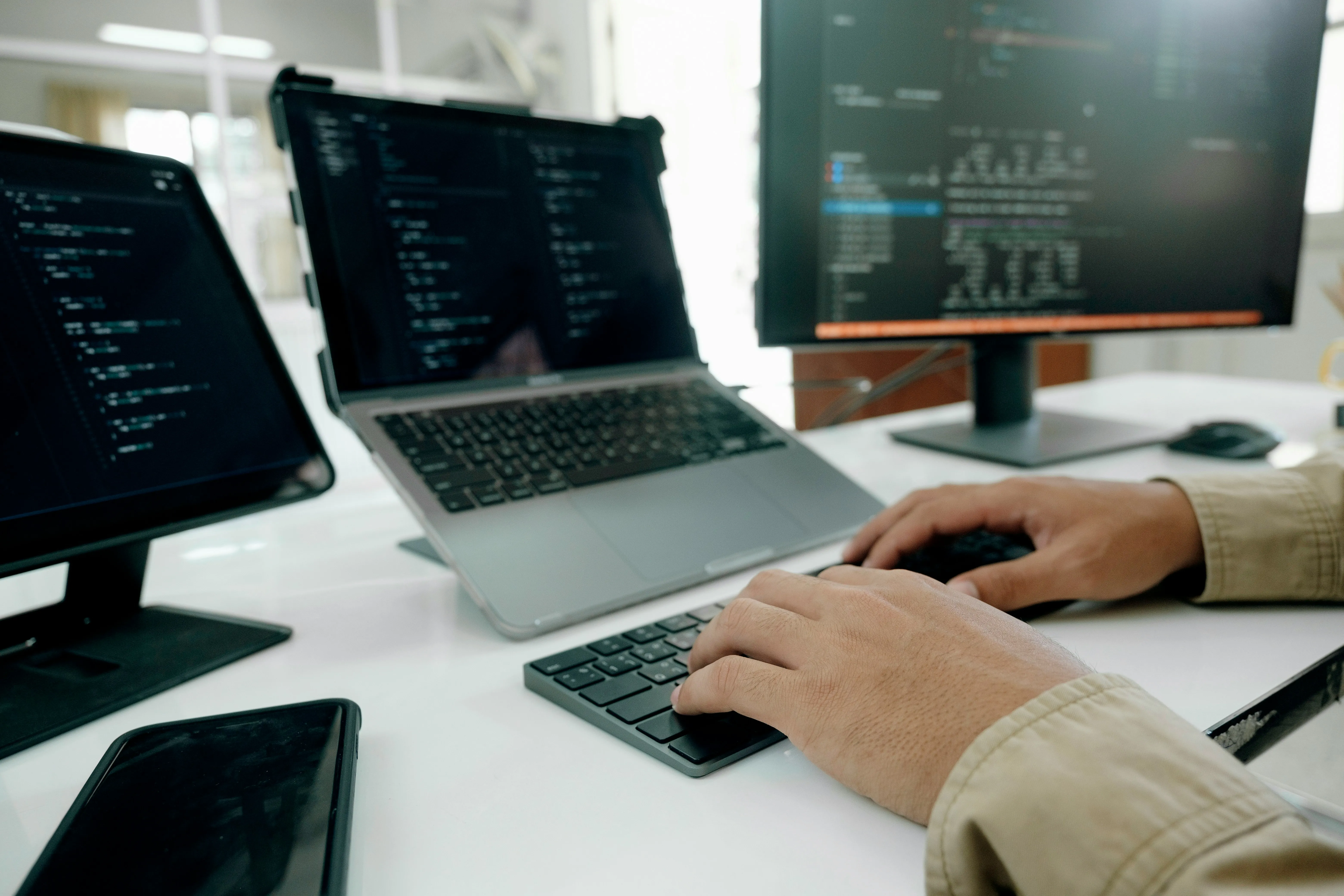 Person typing on a wireless keyboard with code displayed on screens of a laptop, tablet, and monitor in a bright office.