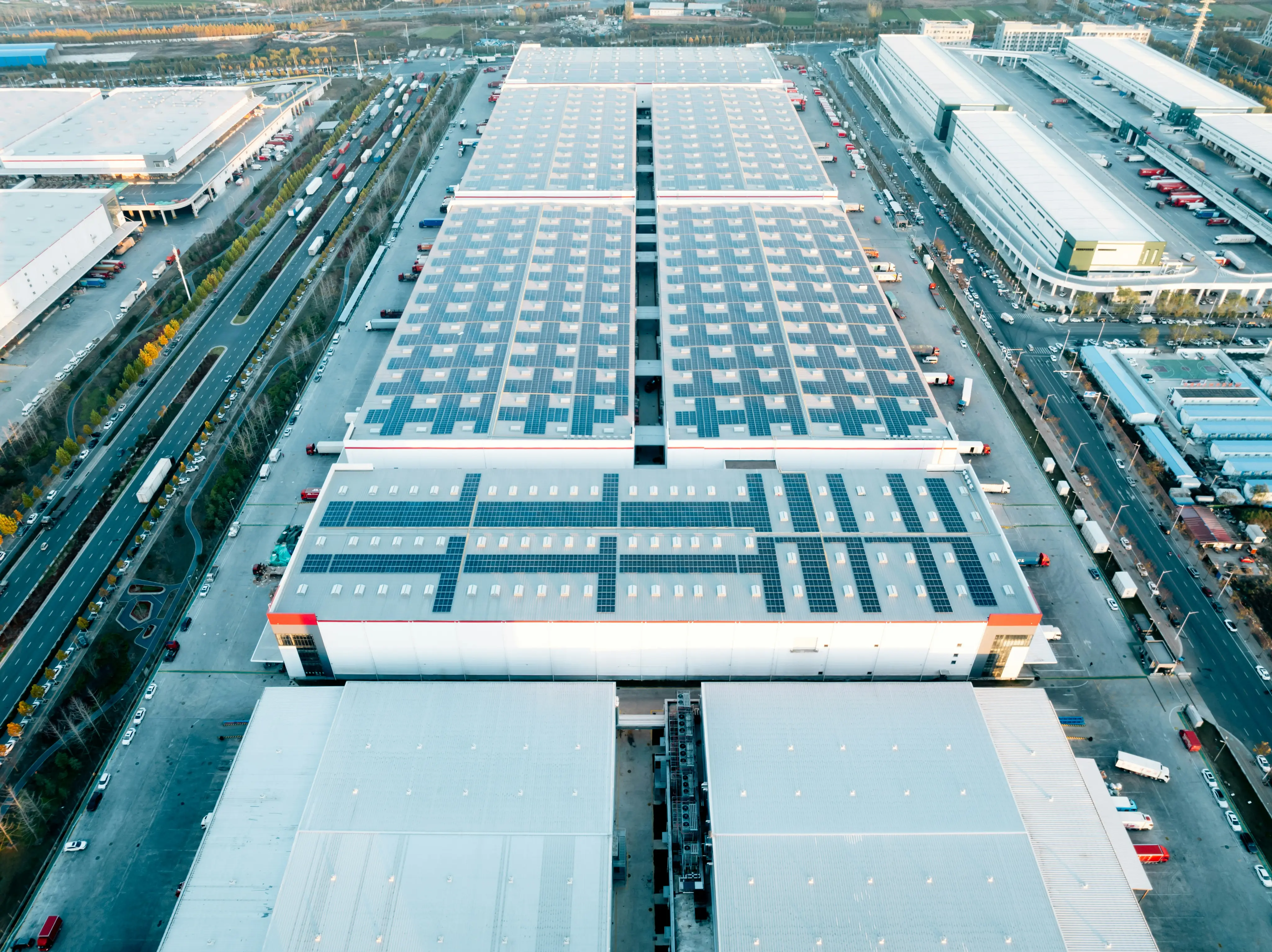 Aerial view of large industrial warehouses with solar panels installed on the roofs and trucks parked around the buildings.