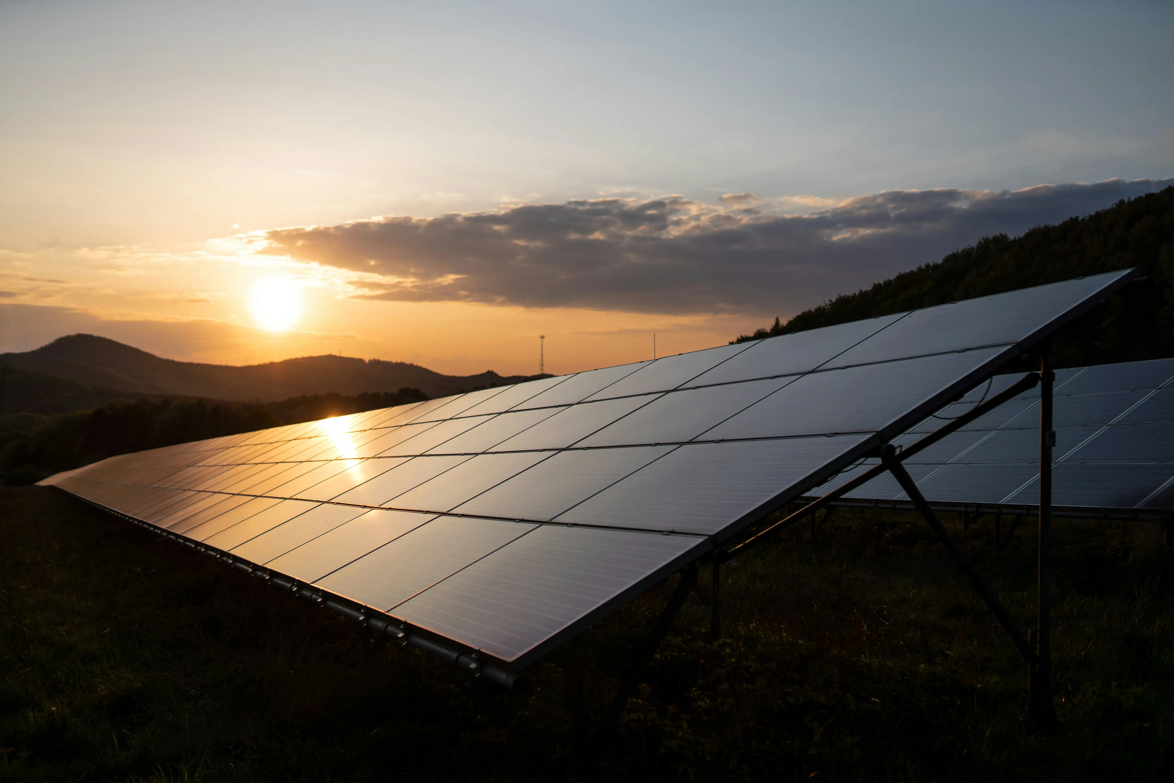 Solar panels angled towards the setting sun on a hillside with scattered clouds and distant mountains.