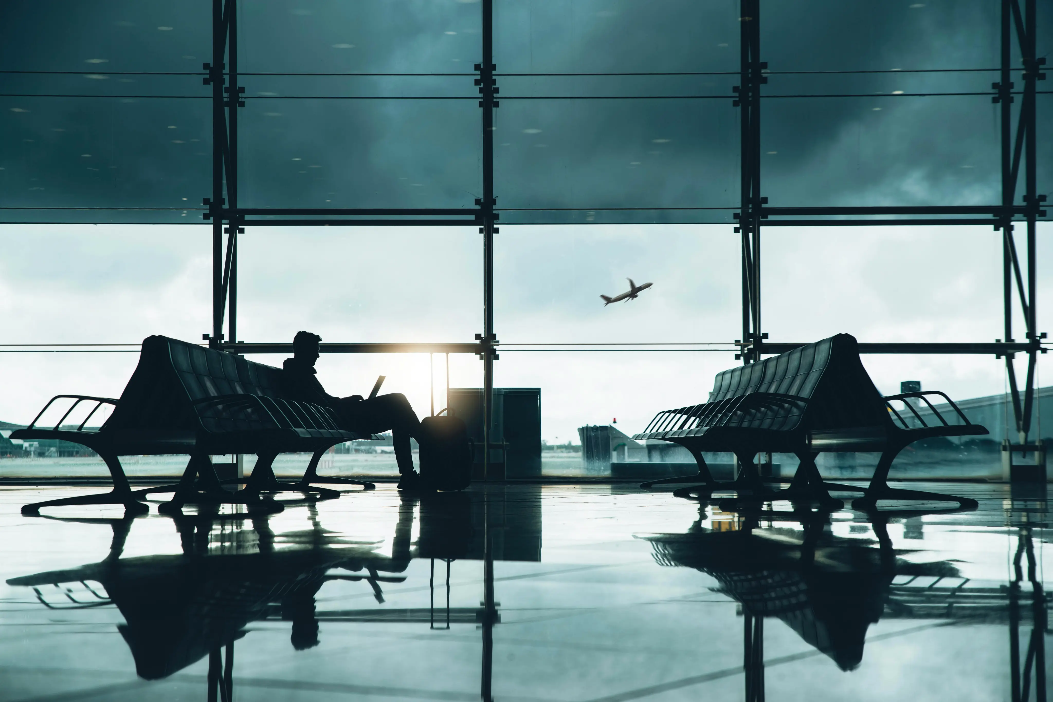 Silhouette of a person sitting with luggage and using a laptop inside an airport terminal, with a plane flying outside the large windows.