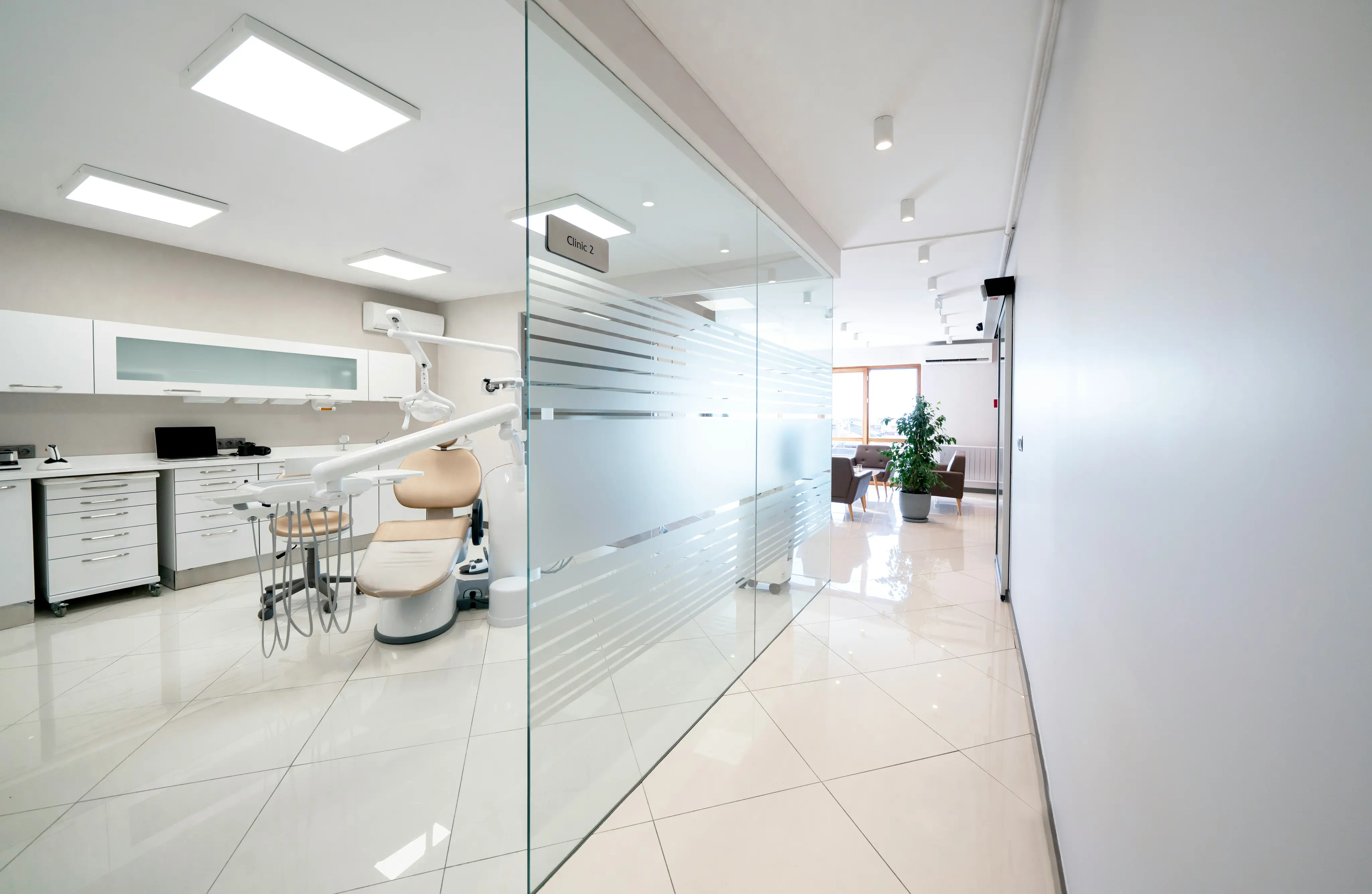 Bright modern dental clinic room with beige dental chair, white cabinets, equipment, and frosted glass partition labelled Clinic 2.
