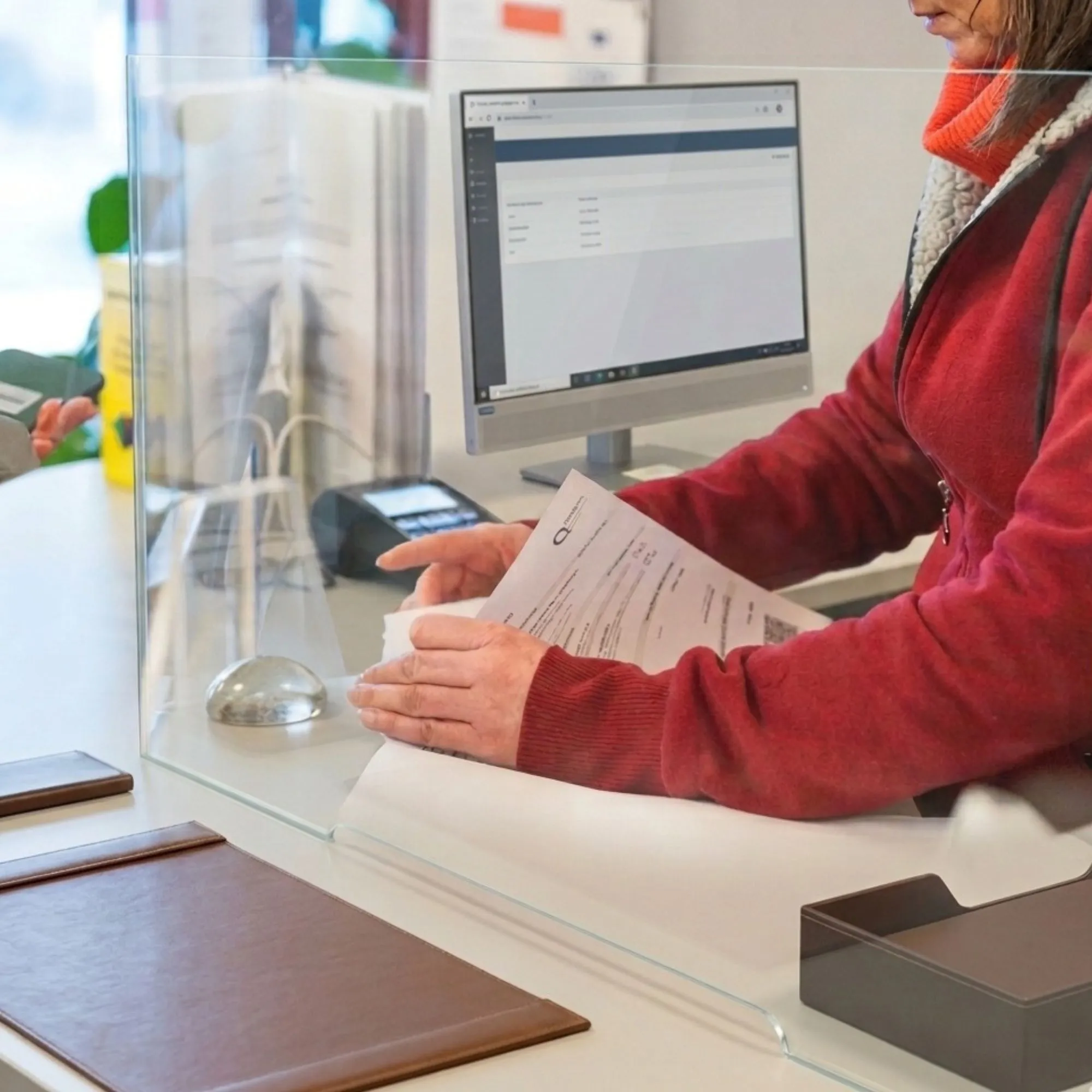 Two women at a service counter, one holding documents and the other holding a wallet, separated by a transparent protective barrier.