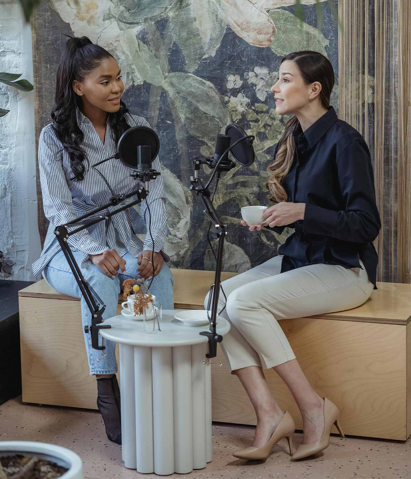 Two women sitting on a bench in front of a large floral artwork, talking into microphones with one holding a coffee cup.
