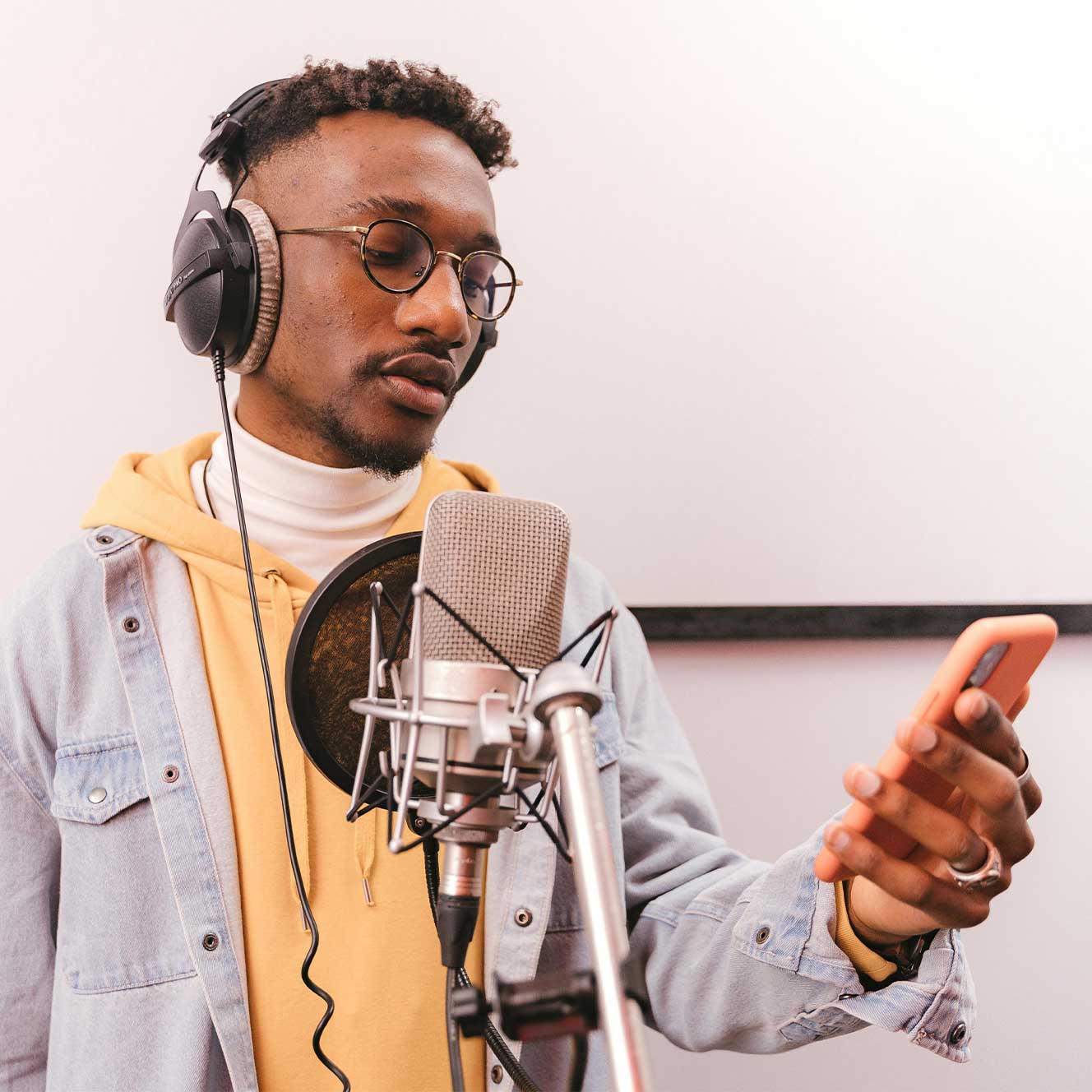Young man wearing headphones and glasses, singing into a studio microphone while holding a smartphone.