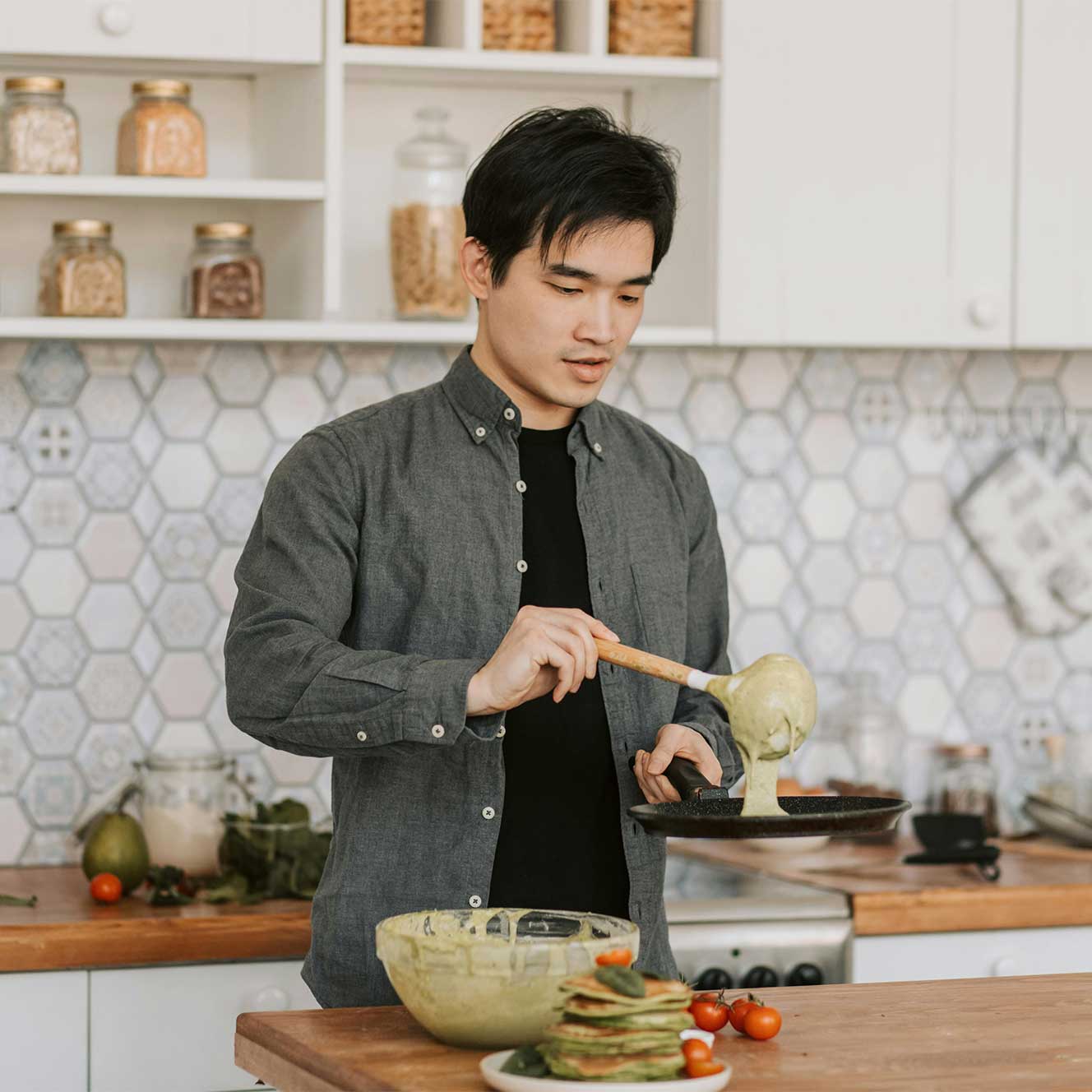 Man pouring green batter from a ladle into a frying pan in a kitchen with a plate of green pancakes and cherry tomatoes on the counter.