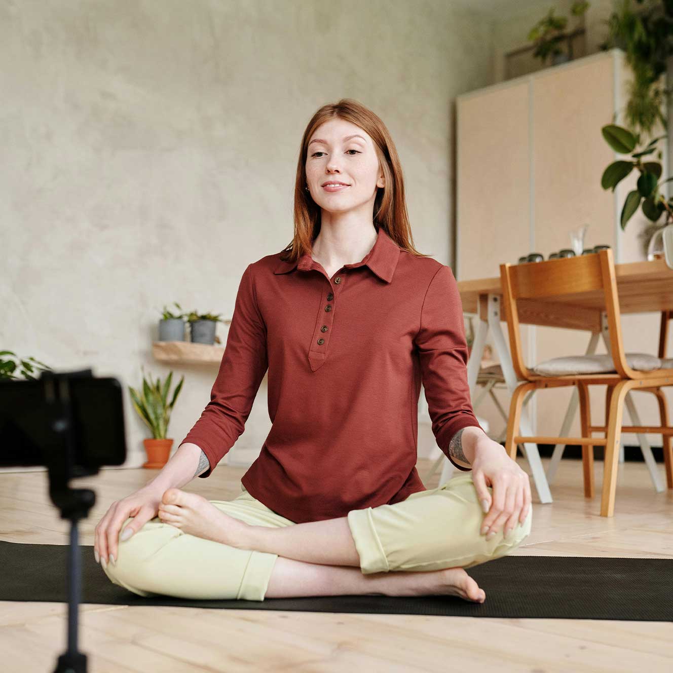 Young woman in a red shirt sitting cross-legged on a yoga mat filming a wellness video indoors.