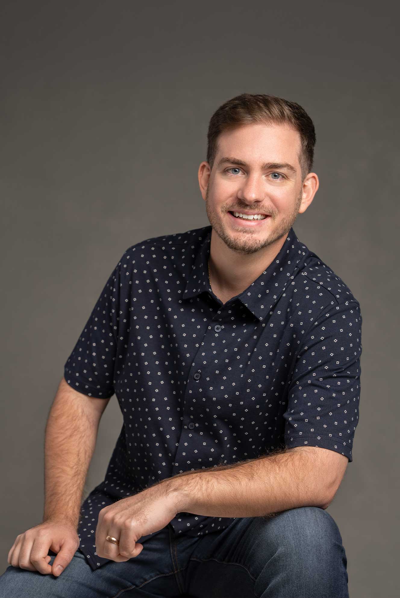 Smiling man with short brown hair and beard wearing a navy blue patterned short-sleeve shirt and jeans, seated against a neutral gray background.