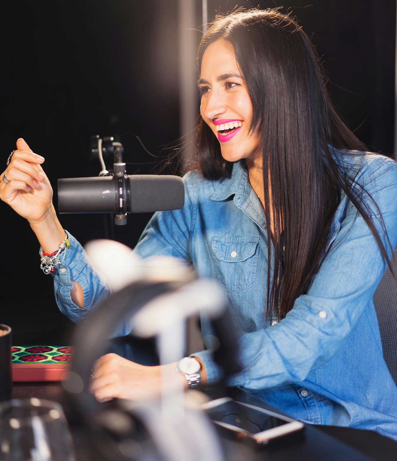 Smiling woman in a denim shirt speaking into a microphone in a podcast recording studio.