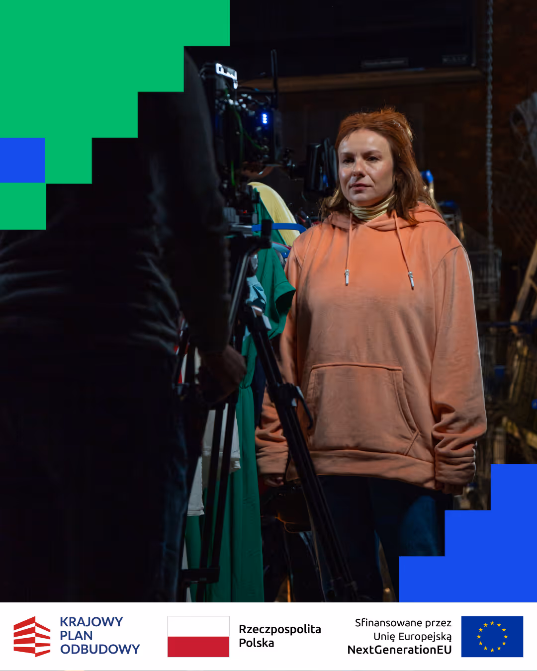 Kobieta w różowej bluzie z kapturem stoi przed kamerą na planie filmowym.