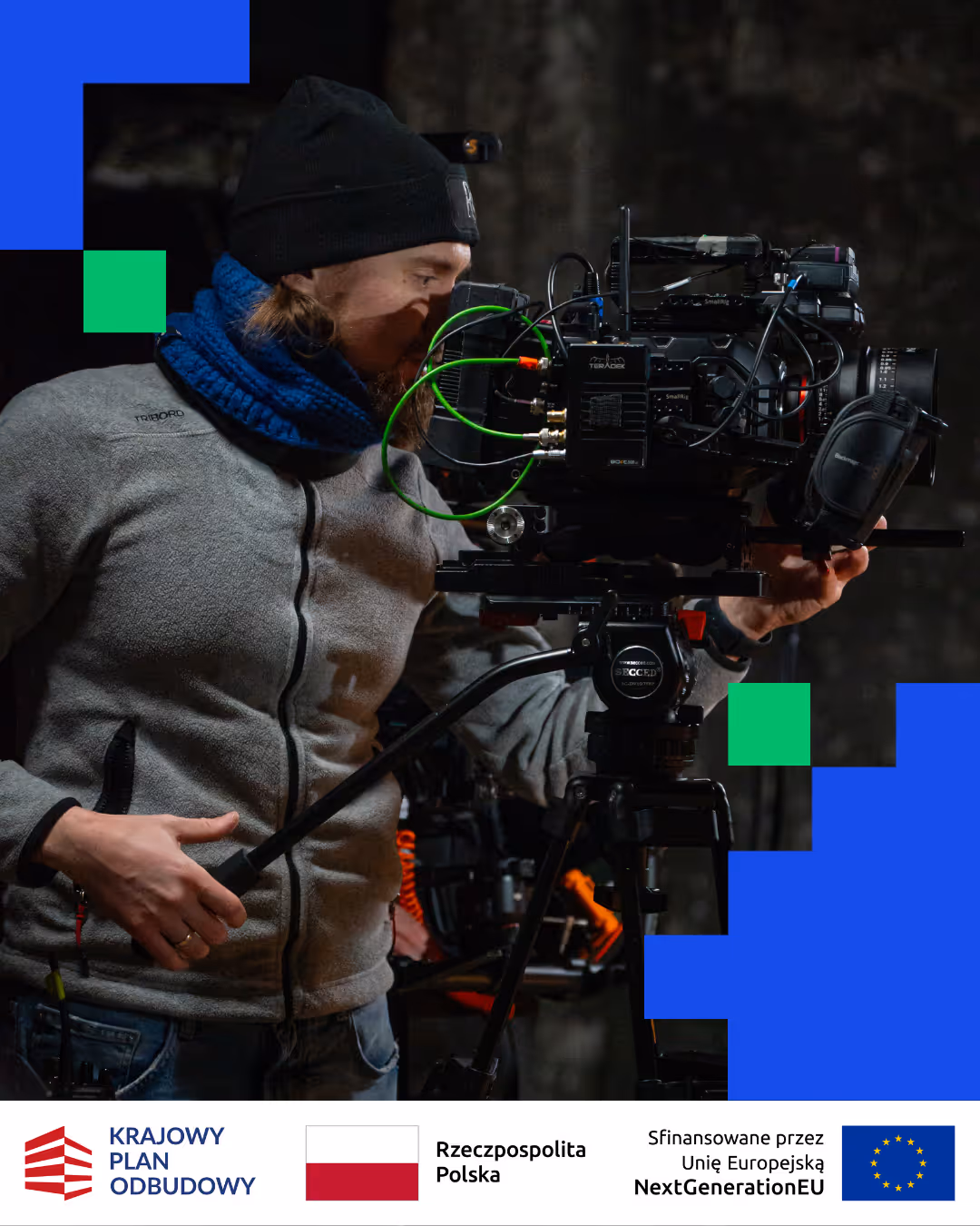 Operator kamery filmowej z czarną czapką i niebieskim szalikiem obsługujący profesjonalną kamerę na statywie w ciemnym otoczeniu.
