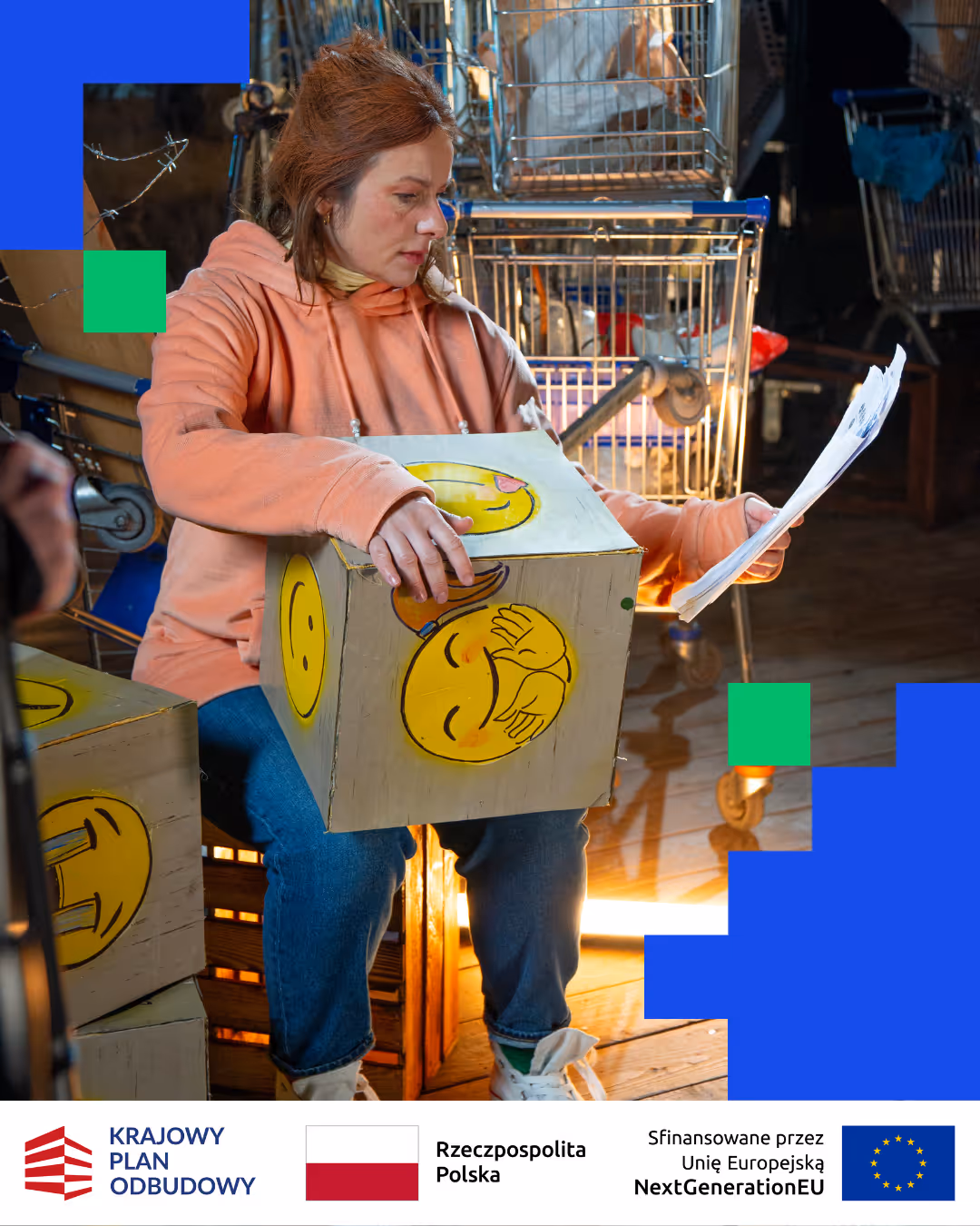 Kobieta w różowej bluzie z kapturem siedzi, trzyma karton ozdobiony emoji, przegląda dokumenty w magazynie z wózkami na zakupy.