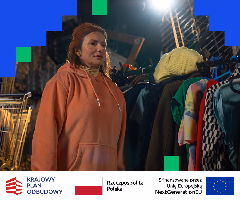 Kobieta w pomarańczowej bluzie z kapturem stoi obok wieszaka z kolorowymi ubraniami i żółtą czapką.