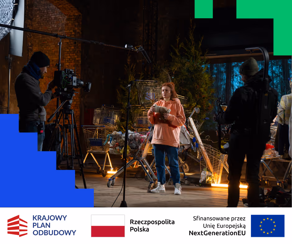 Młoda kobieta w pomarańczowej bluzie stoi przed kamerą na planie filmowym z koszykami sklepowymi i sprzętem filmowym w tle.