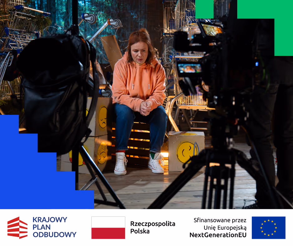 Kobieta w pomarańczowej bluzie siedzi między reklamówkami z emotikonami podczas kręcenia filmu na planie z wózkami sklepowymi w tle.