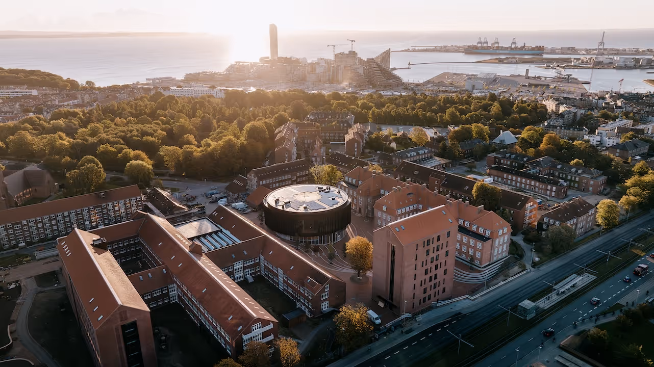Aerial view of urban buildings with red roofs, lush green trees, and a port with cranes by the water at sunset.