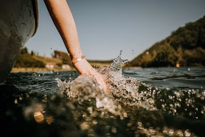 Person's hand splashing water while touching the surface of a lake with blurred hills in the background.