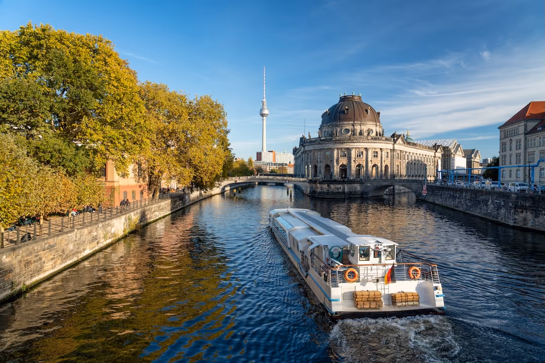 Tour boat cruising on a river in Berlin with the Bode Museum and Berlin TV Tower in the background under a clear blue sky.