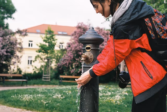 Person in a red jacket filling hands with water from an outdoor decorative water fountain in a park.
