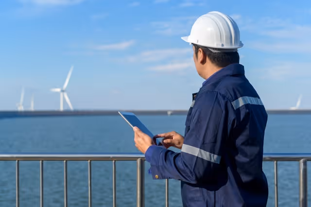 Engineer in safety helmet and blue jacket using a tablet by railing overlooking wind turbines over water.