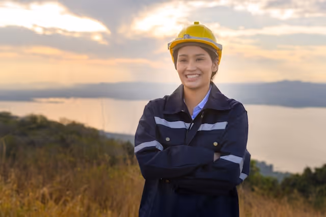 Smiling woman in a yellow hard hat and dark reflective safety jacket standing outdoors with a scenic lake and hills in the background at sunset.