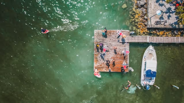 Aerial view of people on a wooden dock and paddleboarding near a white boat in green water.