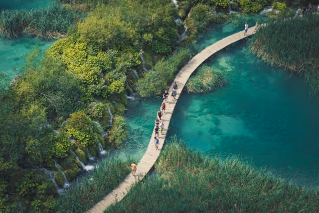 Aerial view of people walking on a curved wooden boardwalk over clear turquoise water surrounded by lush green vegetation.