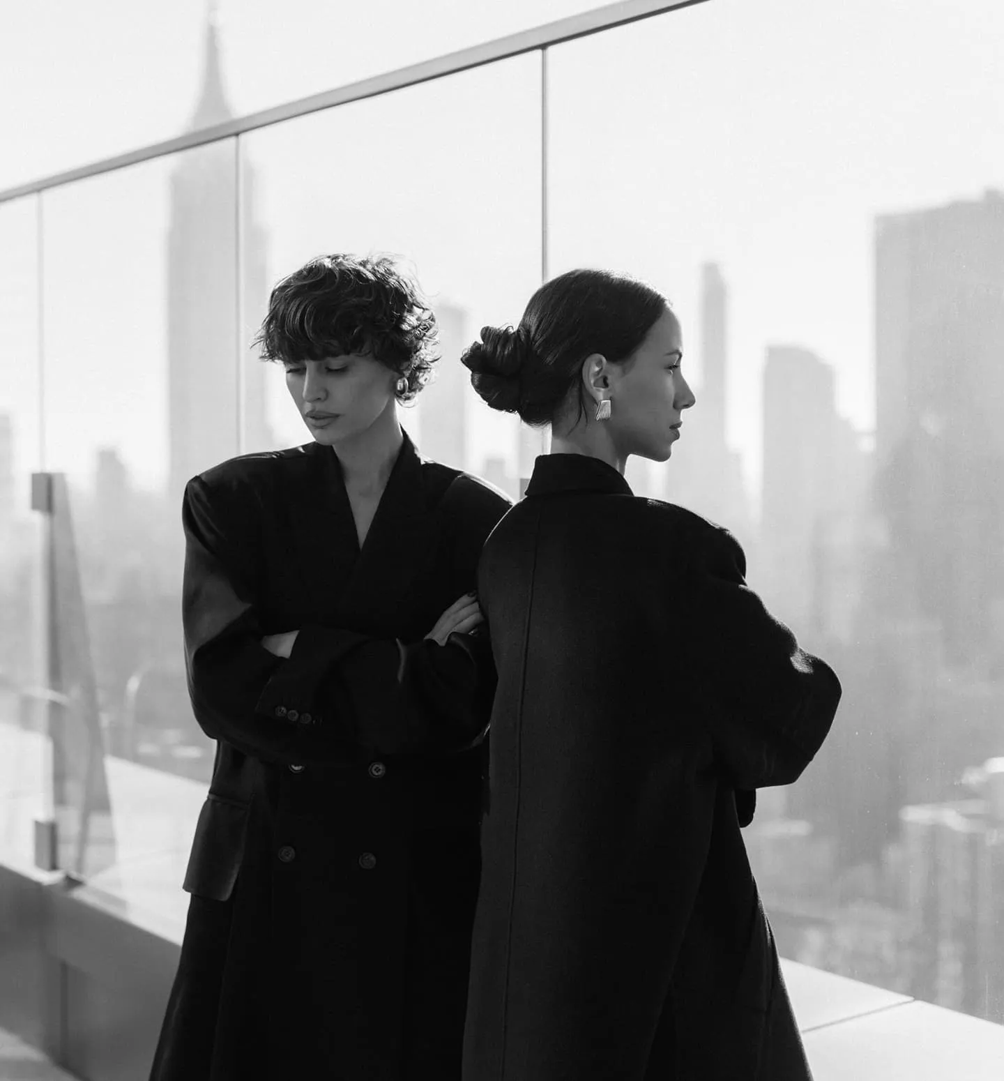 Two women in elegant black coats standing back to back against a modern city skyline, creative agency style.