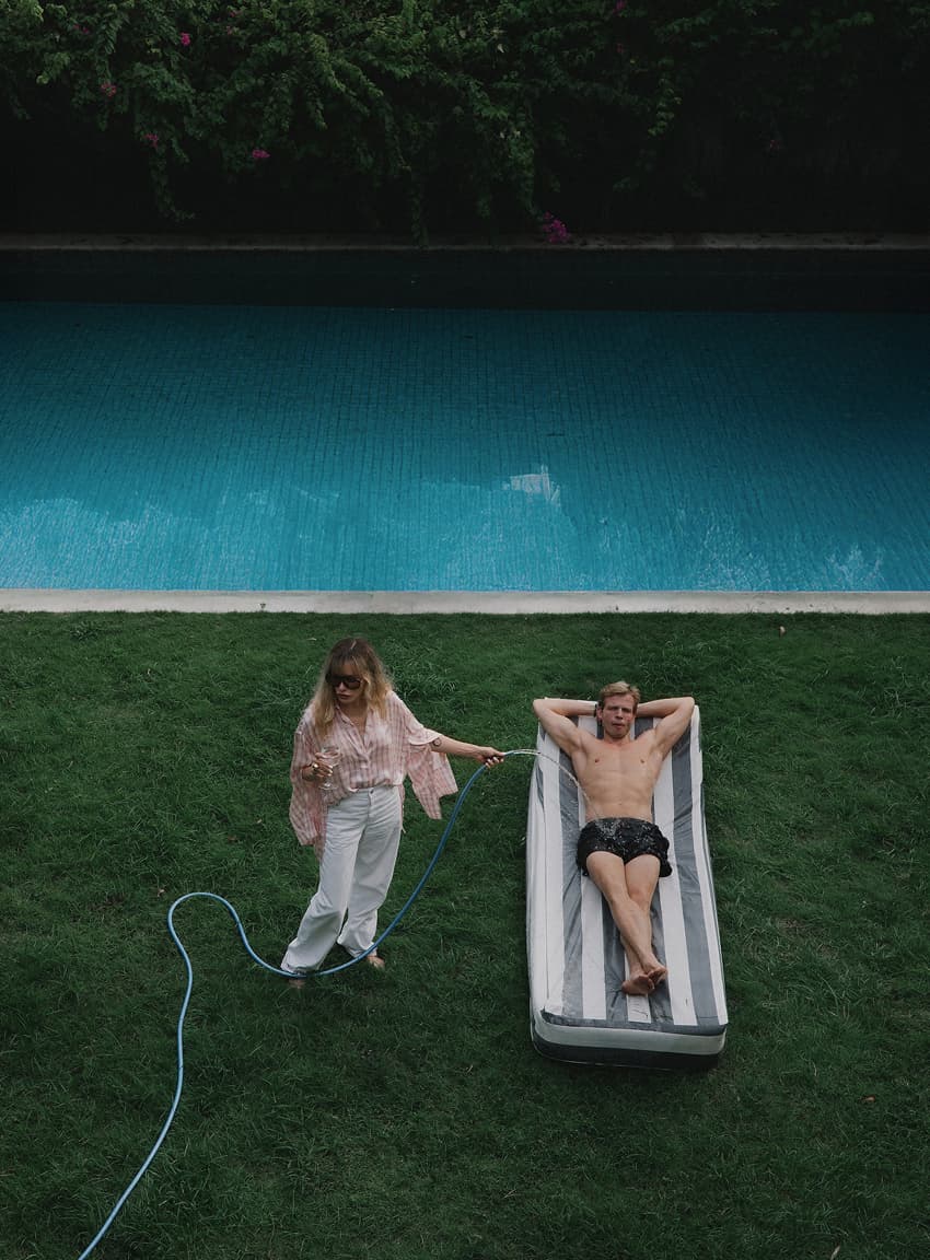 Man relaxing on an inflatable lounger by a pool while a woman waters the lawn, editorial photography for Natalya Pavchinskaya.
