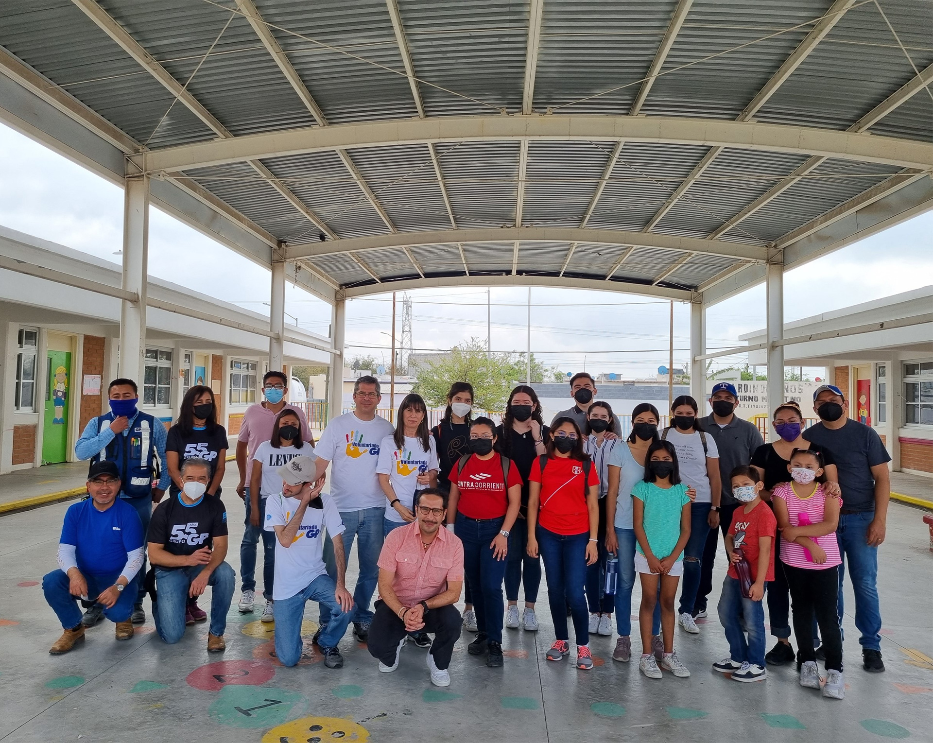 Volunteers and families wearing face masks pose under a roof in a schoolyard.