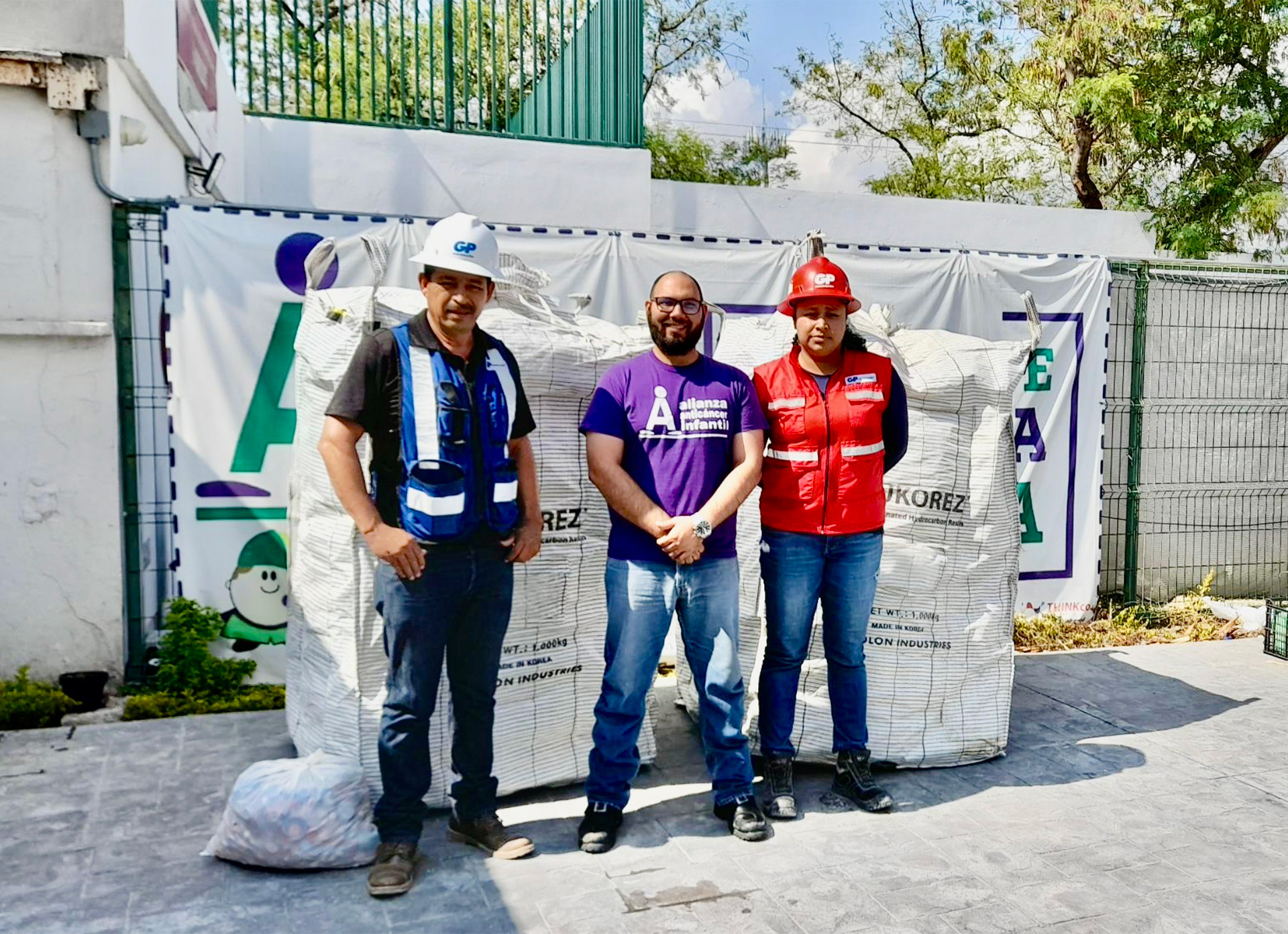 Three people are posing in front of sacks of stored grain, wearing GP vests and a purple T-shirt.