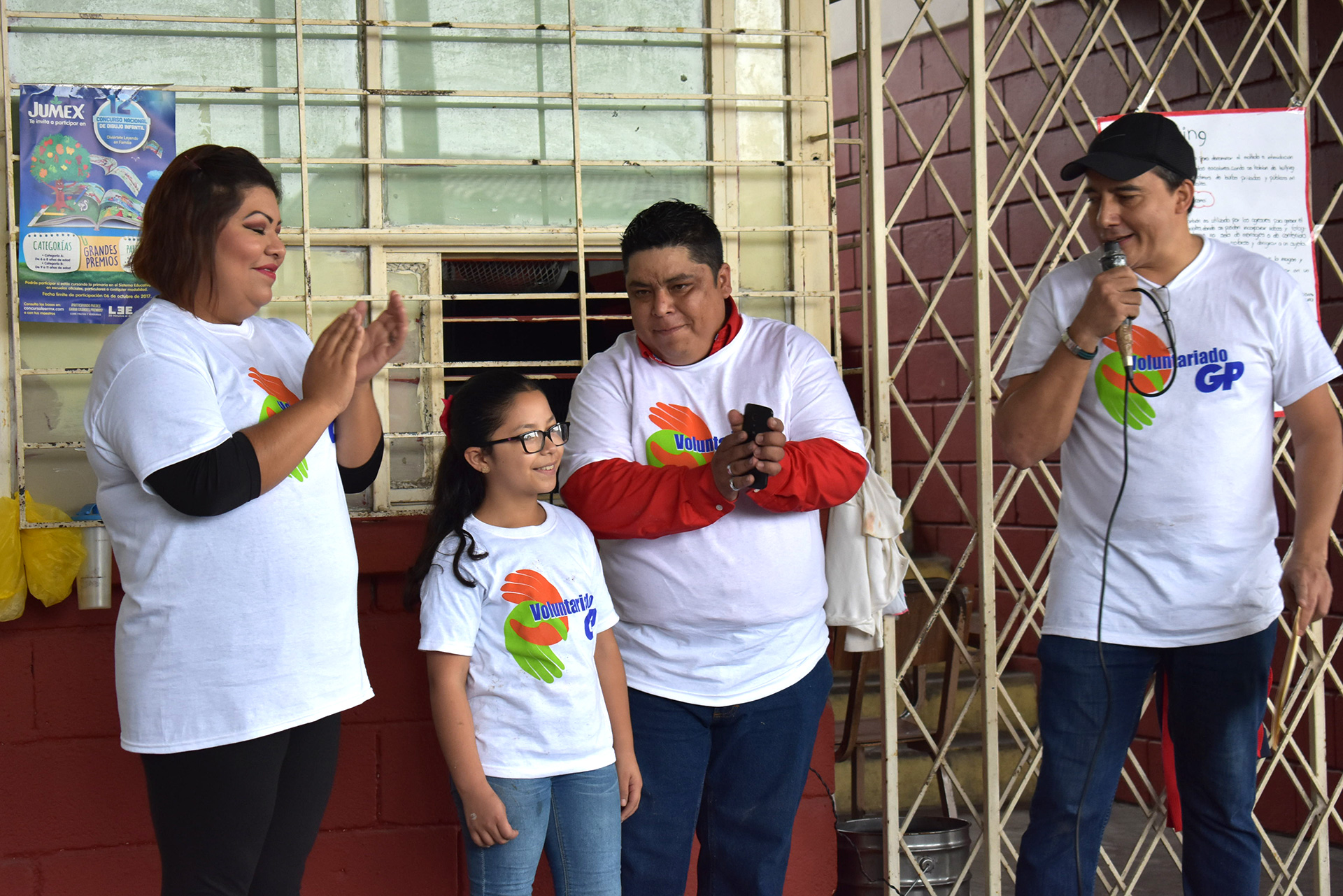 GP volunteers and a little girl are clapping; one of them is speaking into a microphone next to a fence.