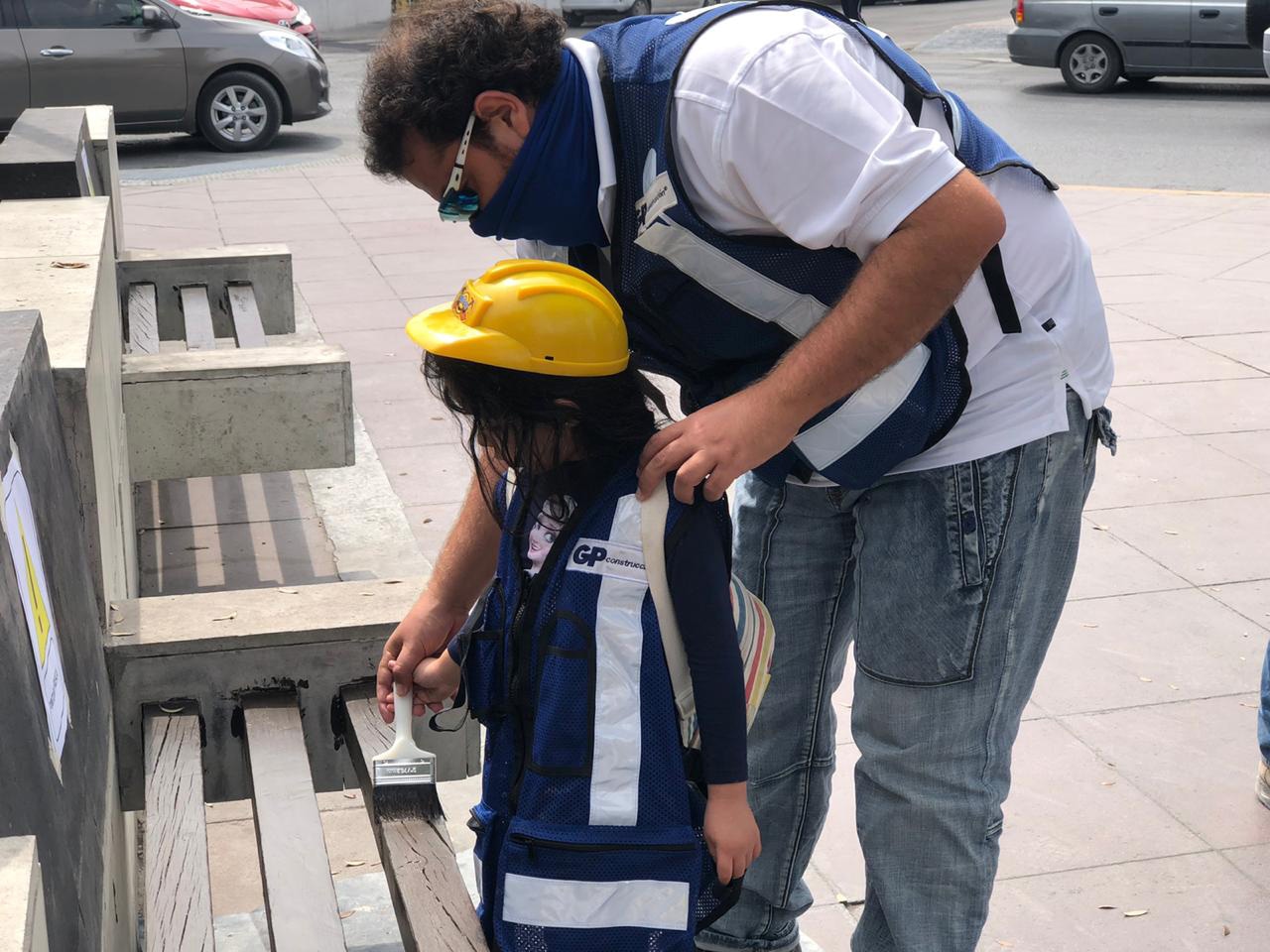 A volunteer adjusts a little girl's yellow vest and helmet on the sidewalk.