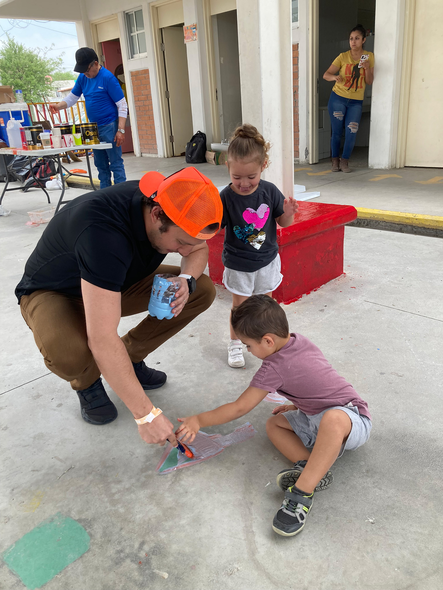 A volunteer and two children are painting on the floor of a hallway, next to a red bench.