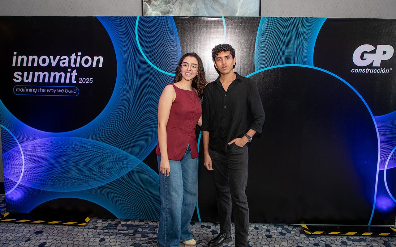 Dos personas jóvenes, una mujer con blusa roja sin mangas y pantalones de mezclilla azules, y un hombre con camisa negra y pantalones oscuros, posan frente a un fondo negro con círculos azules y el texto 'innovation summit 2025 redefining the way we build' y el logo de GP construcción.