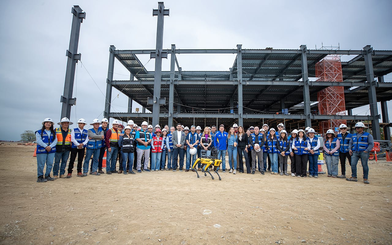 Grupo de trabajadores de construcción usando cascos y chalecos de seguridad, posando frente a una estructura metálica en construcción con un robot cuadrúpedo amarillo en el suelo delante de ellos.