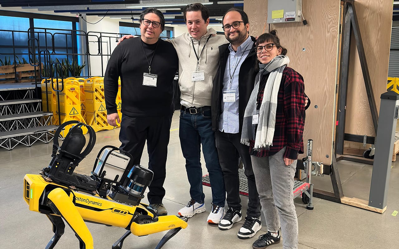 Cuatro personas con gafetes sonríen mientras posan junto a un robot cuadrúpedo amarillo de Boston Dynamics en un espacio industrial con piso de cemento y estructuras metálicas.