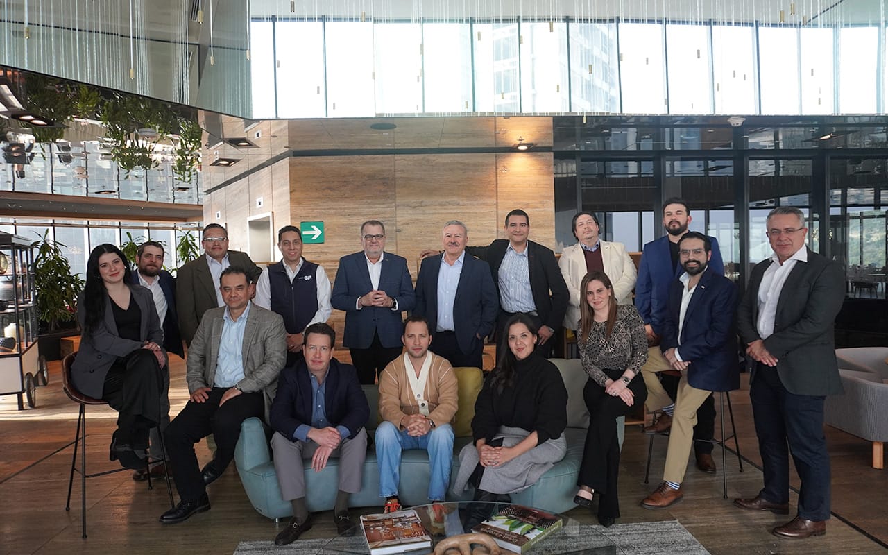 Grupo de quince personas en un entorno de oficina moderno con grandes ventanales, posando para una foto formal, algunos sentados en sofás y otros de pie detrás de ellos.
