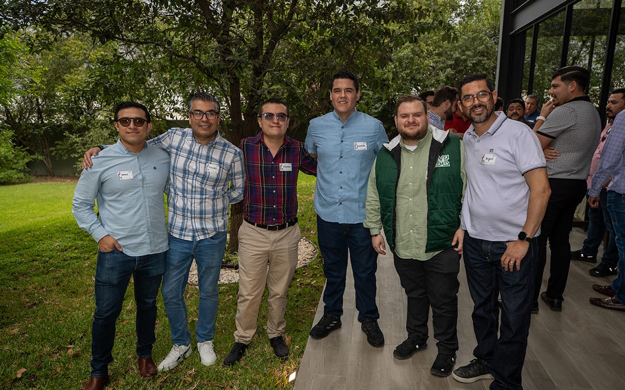 Seis hombres sonrientes posan juntos en un espacio exterior con césped y árboles detrás mientras otros hombres conversan en el fondo cerca de un edificio.