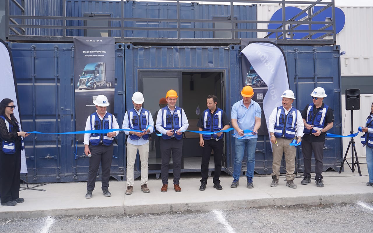 Personas con chalecos azules y cascos blancos y naranjas cortan una cinta azul frente a un recinto hecho con contenedores industriales azules durante un evento de inauguración.