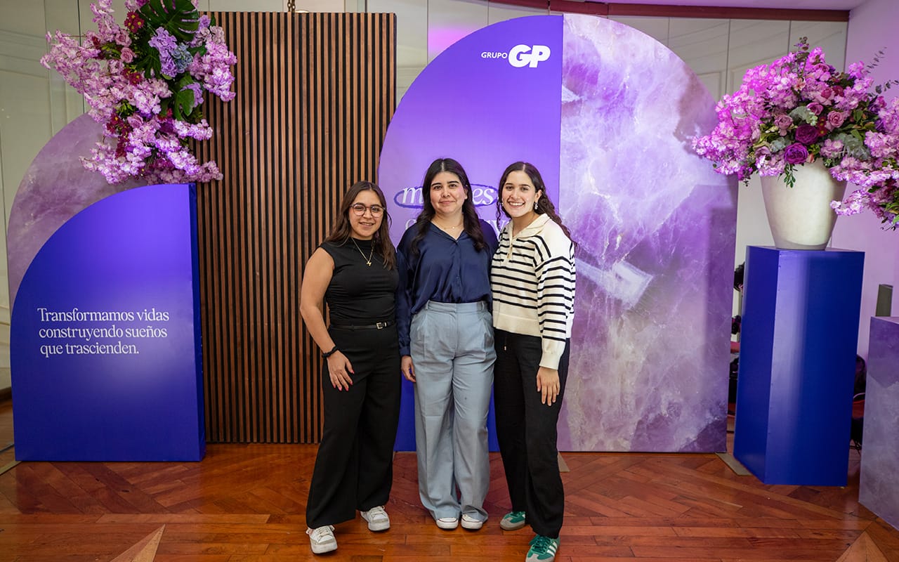 Tres mujeres posan sonrientes en un evento con decoración púrpura que incluye flores y paneles con frases motivacionales de Grupo GP.