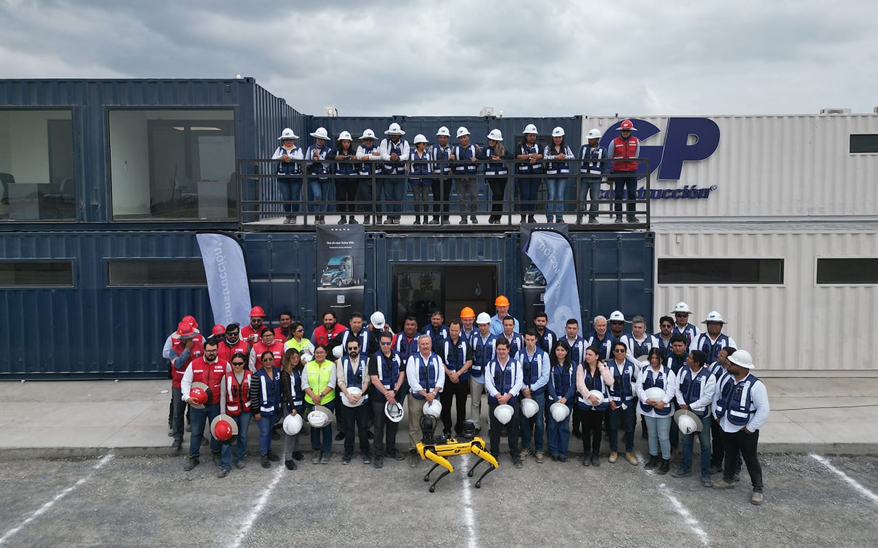 Grupo de trabajadores con cascos y chalecos de seguridad, algunos con cascos rojos y otros blancos, posando frente a una estructura de contenedores azules y blancos con el logo GP Construcción, bajo un cielo nublado. En el suelo, frente al grupo, está un robot cuadrúpedo amarillo.