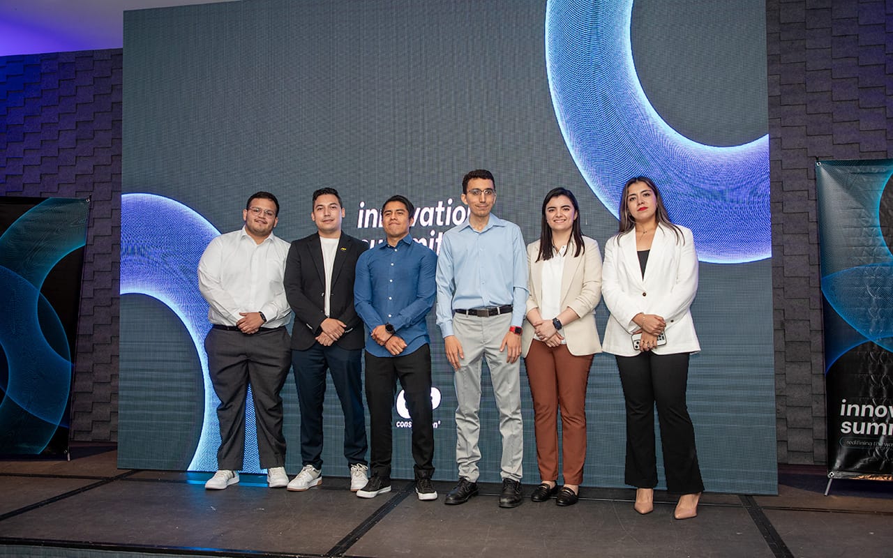 Seis personas están de pie en un escenario frente a una pantalla grande con gráficos circulares azules y texto parcialmente visible que dice 'innovation summit'. Tres hombres y tres mujeres posan, vestidos formalmente en ropa de negocios.